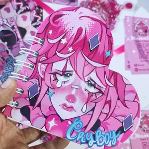 Puede incluir: Un cuaderno en forma de corazón con una ilustración rosa de un personaje llorando, la palabra "Cryboy" en azul. El cuaderno tiene una encuadernación en espiral y está rodeado de otros artículos de papelería rosas.