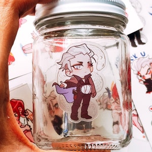 May include: A clear glass jar with a silver lid, held in a hand. Inside the jar is a sticker of a cartoon character with white hair, a purple cape, and brown trousers. Other stickers are visible.