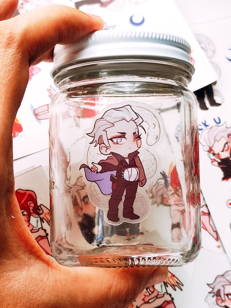 May include: A clear glass jar with a silver lid, held in a hand. Inside the jar is a sticker of a cartoon character with white hair, a purple cape, and brown trousers. Other stickers are visible.