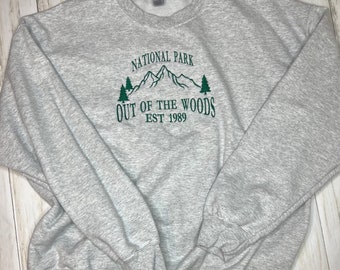 May include: A heather gray sweatshirt with the words "NATIONAL PARK OUT OF THE WOODS EST 1989" in green. The design includes a mountain range and two trees. The sweatshirt has a crew neck and long sleeves.
