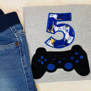 Op de afbeelding: Een grijs T-shirt met een blauw en geel nummer 5 en een zwarte videogamecontroller. Het nummer 5 is versierd met een patroon van videogamecontrollers.