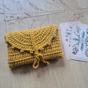 May include: Mustard yellow crocheted clutch with a flap closure and a knotted tie. Two tarot cards with floral and symbolic illustrations are next to the clutch. The clutch and cards are on a light wood surface.