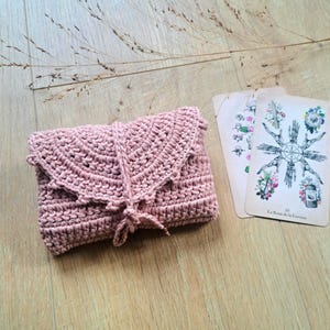 May include: A dusky pink crochet wallet with a flap and tie closure. Two tarot cards with floral and wheel illustrations are next to the wallet. The items are on a light wood surface with dried grass.