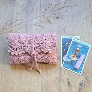 Könnte beinhalten: Ein altrosa gehäkeltes Täschchen mit einer bogenförmigen Klappe und einem Bindeband mit kleinen Holzperlen. Das Täschchen liegt neben zwei Tarotkarten, La Maison-Dieu und As des Coupes, sowie einem klaren Kristallstab.