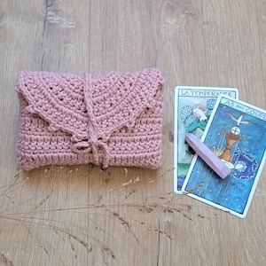 May include: A dusky pink crochet clutch bag with a flap and wooden bead detail. Two tarot cards and a pink crystal are next to the bag. The cards have French text.