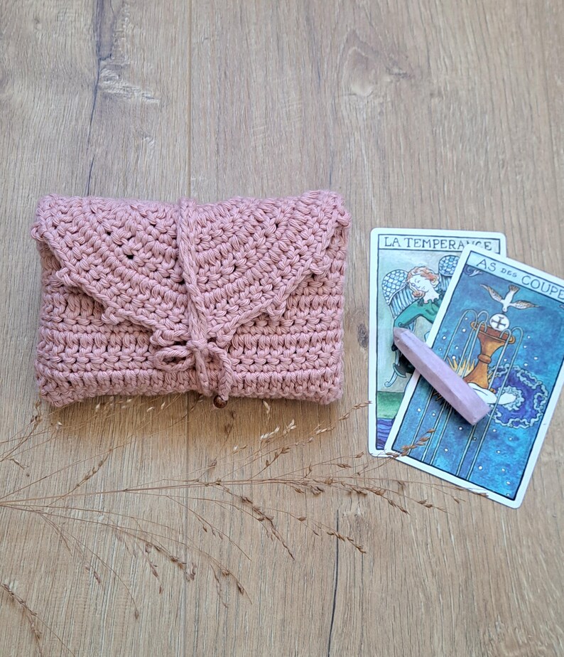 May include: A dusky pink crochet clutch bag with a flap and wooden bead detail. Two tarot cards and a pink crystal are next to the bag. The cards have French text.