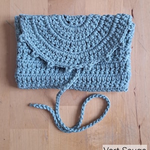 May include: A sage green crocheted clutch bag with a flap and drawstring closure. The handmade bag is made of a thick, textured yarn. The text "Vert Sauge" is visible in the bottom right corner.
