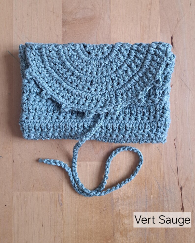 May include: A sage green crocheted clutch bag with a flap and drawstring closure. The handmade bag is made of a thick, textured yarn. The text "Vert Sauge" is visible in the bottom right corner.