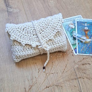 May include: A cream-coloured, crocheted pouch with a flap closure and a drawstring. The pouch is rectangular and has a textured appearance. Two tarot cards are visible in the background. The cards have illustrations and text.