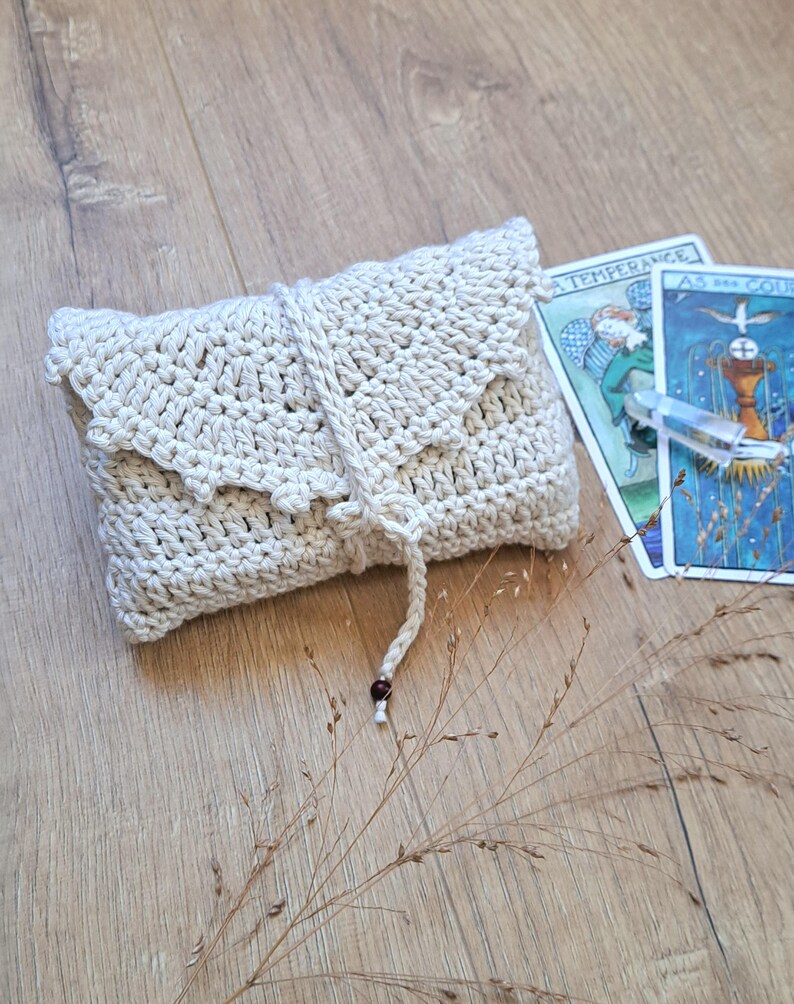 May include: A cream-coloured, crocheted pouch with a flap closure and a drawstring. The pouch is rectangular and has a textured appearance. Two tarot cards are visible in the background. The cards have illustrations and text.