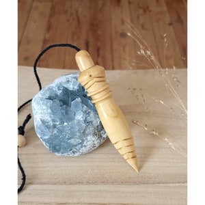 May include: A wooden pendulum with a pointed tip and a string attached. The pendulum is resting on a grey crystal rock.