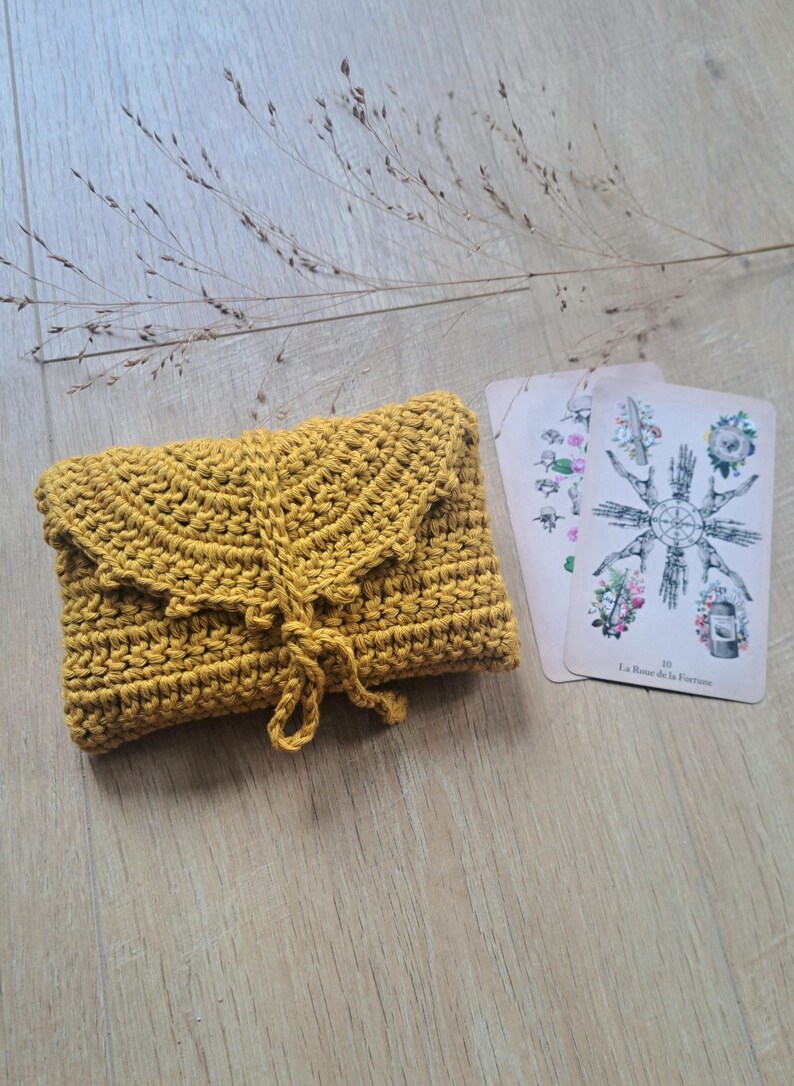 May include: Mustard yellow crocheted clutch with a flap closure and a knotted tie. Two tarot cards with floral and symbolic illustrations are next to the clutch. The clutch and cards are on a light wood surface.