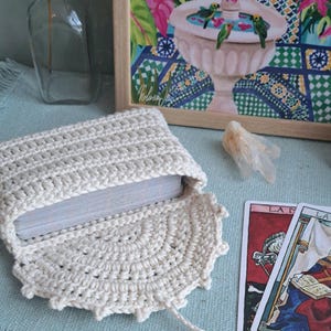 May include: A cream-coloured, open crochet tarot card pouch with a deck of cards inside. The pouch is on a blue surface with a fringed edge. A framed painting with a fountain and parrots is in the background.