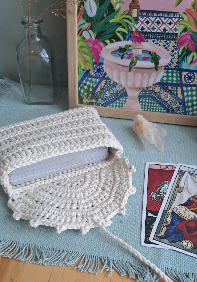 May include: A cream-coloured, open crochet tarot card pouch with a deck of cards inside. The pouch is on a blue surface with a fringed edge. A framed painting with a fountain and parrots is in the background.