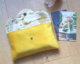 May include: A yellow fabric pouch with a scalloped flap featuring a gold metallic print of a crescent moon, sun, and stars. The pouch has a metal snap closure. Beside it lies a clear quartz crystal cluster and a tarot card labelled Page of Cups.