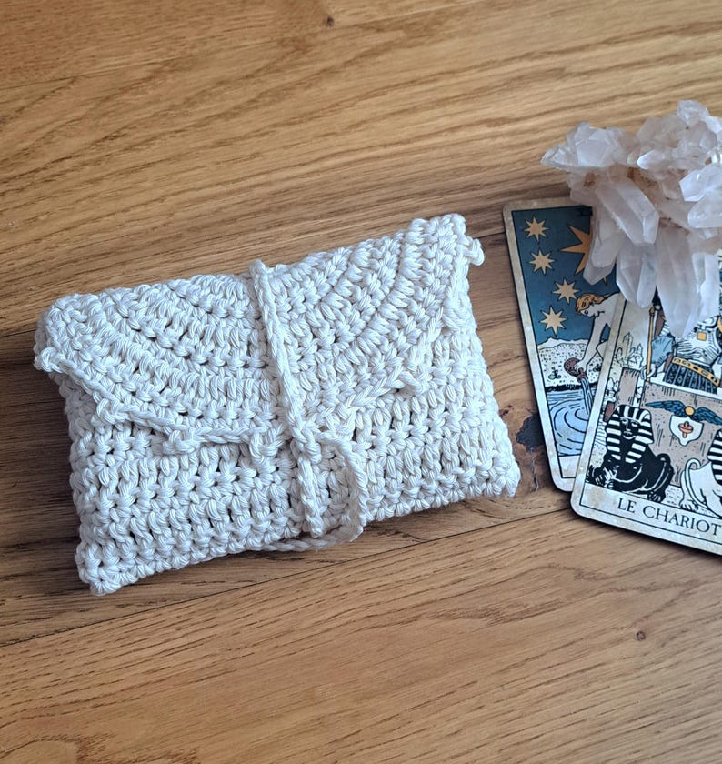 May include: A cream-coloured, crocheted envelope-style pouch with a flap closure and a tie. The pouch is lying on a wooden surface next to tarot cards and a cluster of clear crystals. The tarot cards have illustrations and the words "LE CHARIOT".