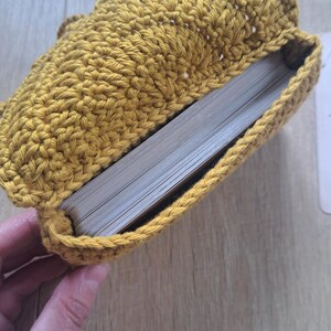 May include: Mustard yellow crochet book sleeve, open to reveal the pages of a book. The handmade sleeve is crafted with a textured stitch, and the book is partially visible. The sleeve is designed to protect books and is a cozy accessory.
