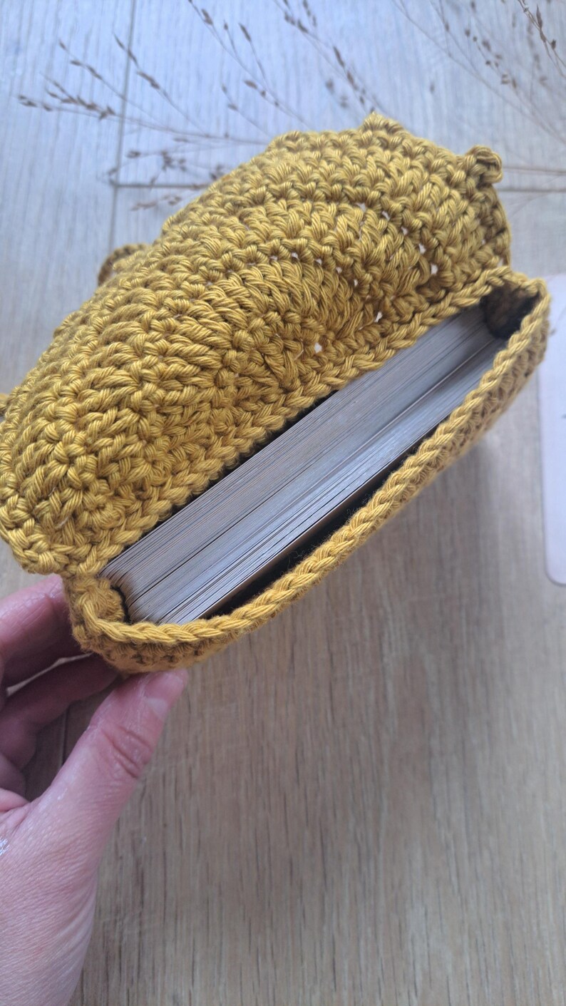 May include: Mustard yellow crochet book sleeve, open to reveal the pages of a book. The handmade sleeve is crafted with a textured stitch, and the book is partially visible. The sleeve is designed to protect books and is a cozy accessory.