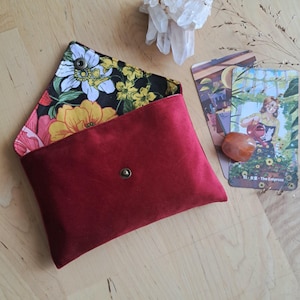 Op de afbeelding: Een rechthoekig envelop-achtig tasje gemaakt van dieprode fluwelen stof. De binnenvoering heeft een levendig bloemenpatroon met gele, witte en roze bloemen op een zwarte achtergrond. Een enkele metalen drukknoop sluit de klep af.