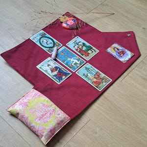 Op de afbeelding: Een dieprood fluwelen tarotkleed met een omslagzak voorzien van een geel en roze bloemrijk toile-patroon. Zes tarotkaarten zijn op het oppervlak gerangschikt, vergezeld door een kleine roze kristal en een decoratief beschilderd hartornament.