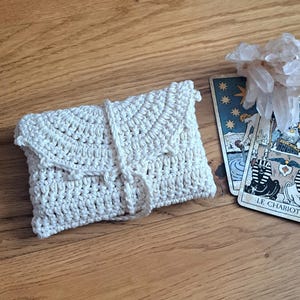 May include: A cream-coloured, crocheted envelope-style pouch with a flap closure and a tie. The pouch is lying on a wooden surface next to tarot cards and a cluster of clear crystals. The tarot cards have illustrations and the words "LE CHARIOT".