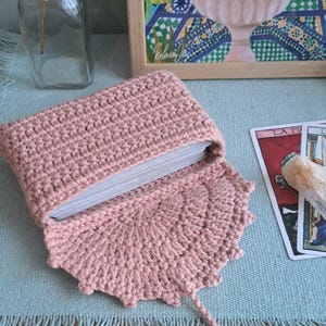 May include: A light pink, crocheted tarot card pouch, open to show a deck of cards. The pouch has a decorative edged flap and a long, braided tie. A framed painting and tarot cards are in the background.
