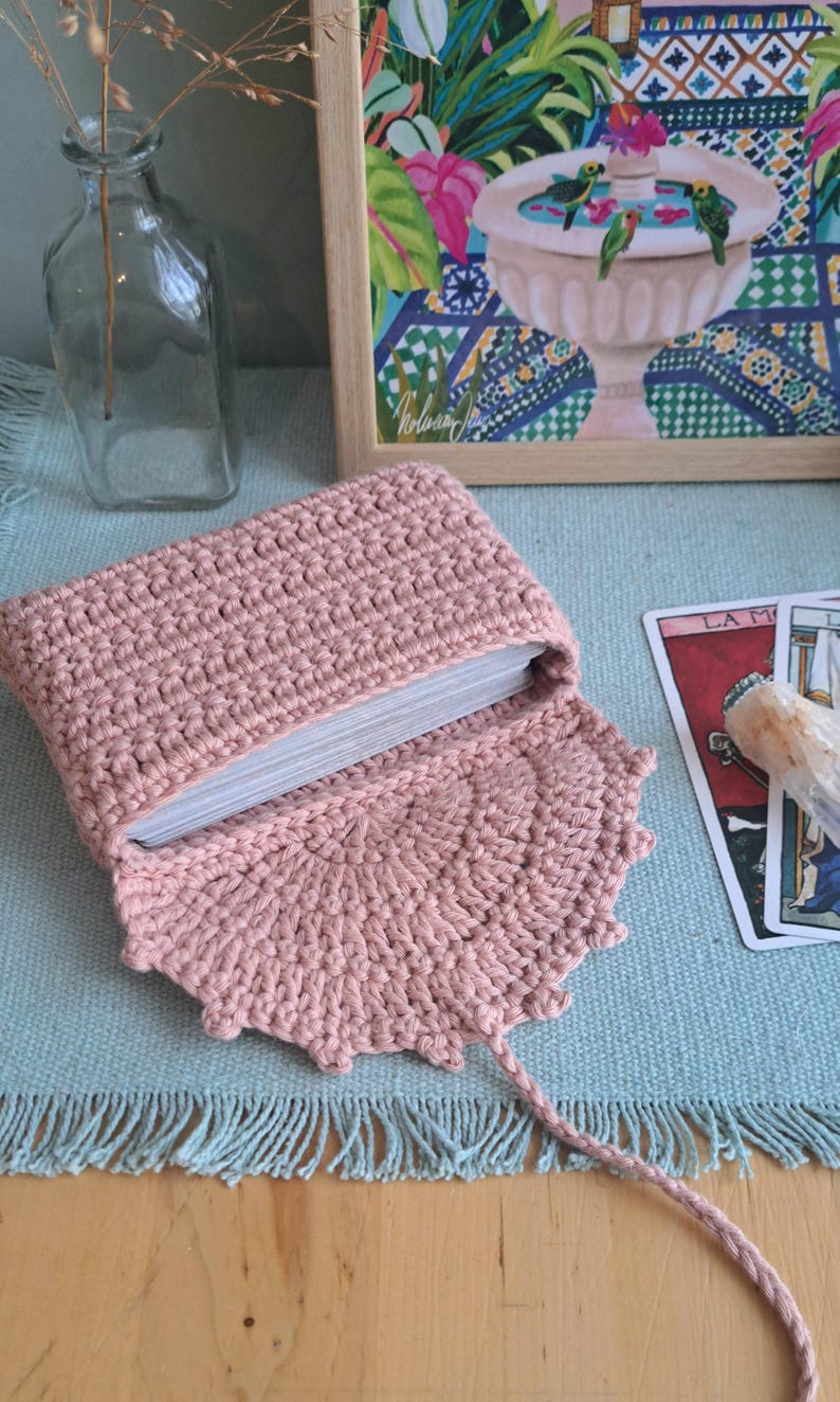 May include: A light pink, crocheted tarot card pouch, open to show a deck of cards. The pouch has a decorative edged flap and a long, braided tie. A framed painting and tarot cards are in the background.