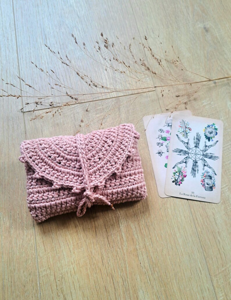 May include: A dusky pink crochet wallet with a flap and tie closure. Two tarot cards with floral and wheel illustrations are next to the wallet. The items are on a light wood surface with dried grass.