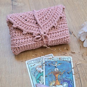 May include: A dusky pink crochet clutch bag with a flap and tie closure. Two tarot cards with illustrations and text are below the bag. A pink crystal and dried plants are also in the image.