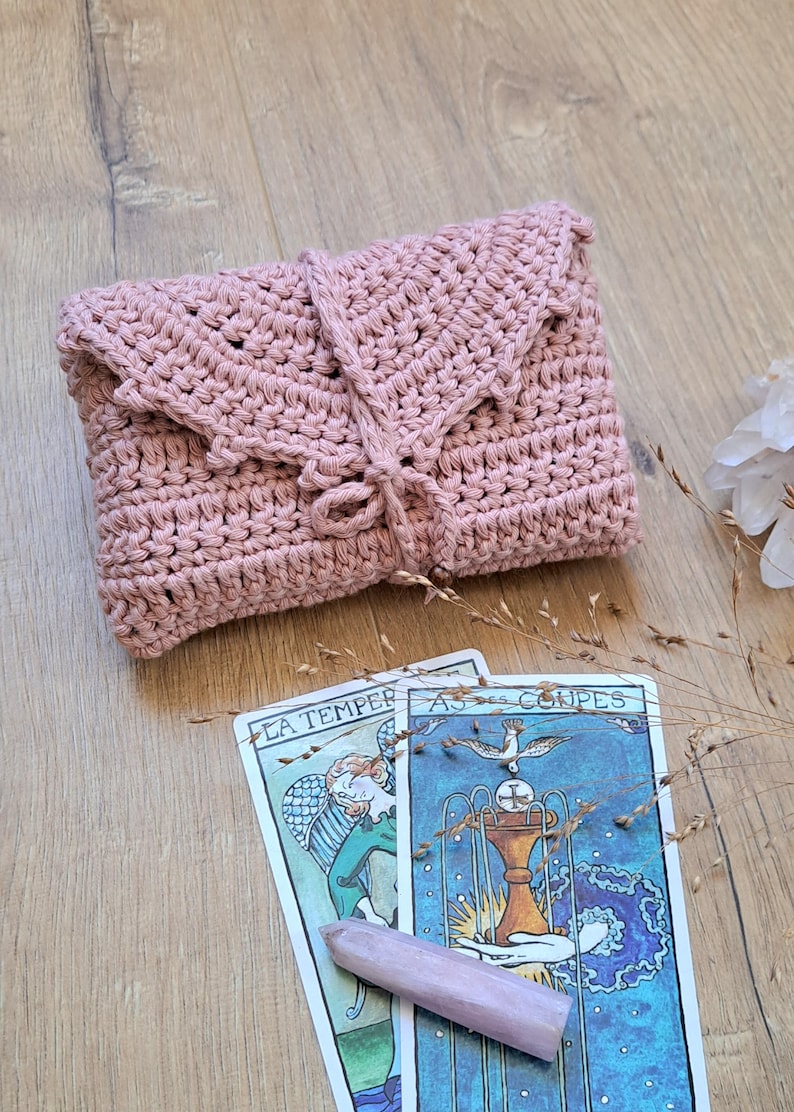 May include: A dusky pink crochet clutch bag with a flap and tie closure. Two tarot cards with illustrations and text are below the bag. A pink crystal and dried plants are also in the image.