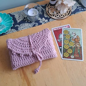 May include: A lilac crochet tarot card pouch with a flap and tie closure. Two tarot cards are displayed, alongside a candle, essential oil bottle, and crystals on a wooden table. The cards are "La Mort" and "Nine of Pentacles."