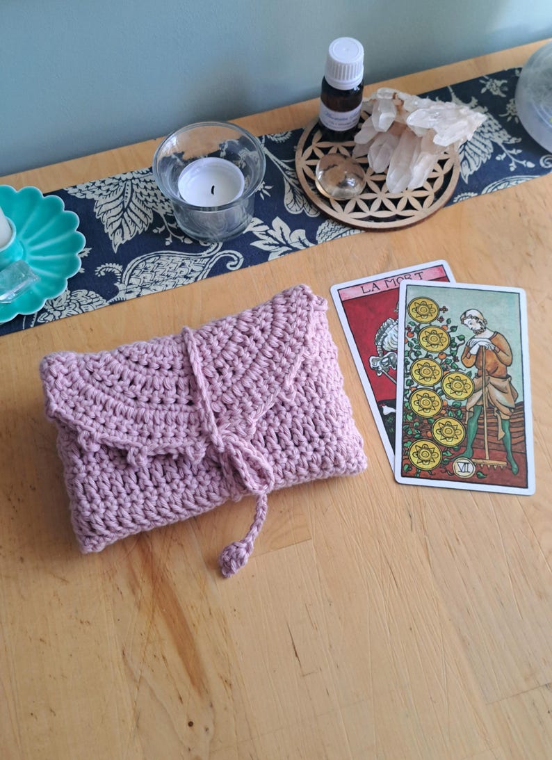 May include: A lilac crochet tarot card pouch with a flap and tie closure. Two tarot cards are displayed, alongside a candle, essential oil bottle, and crystals on a wooden table. The cards are "La Mort" and "Nine of Pentacles."