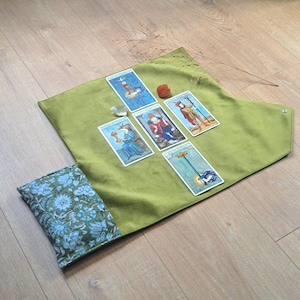 Può includere: Un tappetino per tarocchi in tessuto verde lime con una tasca a motivo floreale blu e bianco su un lato. Cinque carte dei tarocchi sono disposte a croce sul tappetino, accompagnate da un piccolo cristallo trasparente e una pietra arancione liscia.