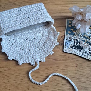 May include: An off-white, crocheted tarot card holder with a decorative scalloped edge. The holder is open, revealing a deck of cards. A tarot card with the text "LE CHARIOT" is visible. The holder is on a wooden surface.