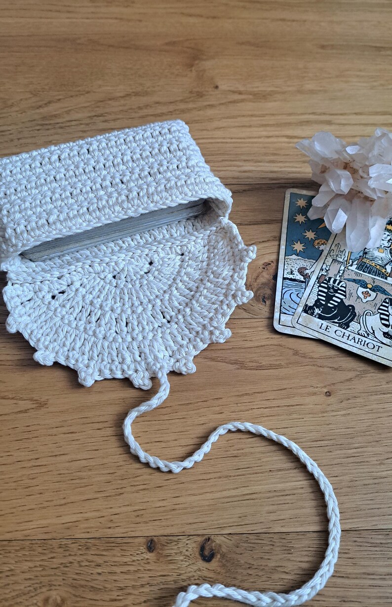 May include: An off-white, crocheted tarot card holder with a decorative scalloped edge. The holder is open, revealing a deck of cards. A tarot card with the text "LE CHARIOT" is visible. The holder is on a wooden surface.