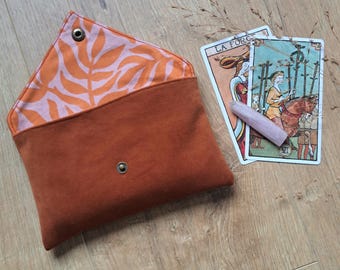 May include: A rust-coloured envelope-style pouch with a pink and orange leaf pattern lining. Two tarot cards and a small, light-coloured wooden object are next to the pouch. The tarot cards have the words "LA FORCE" printed on them.