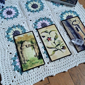 Puede incluir: Una manta blanca de ganchillo con patrones florales en tonos azules, rosas y grises. Se muestran tres cartas de tarot, junto con una baraja de cartas con un emblema de corazón dorado. Las cartas presentan ilustraciones botánicas y arquitectónicas.