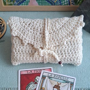 May include: A cream-coloured, hand-crocheted clutch bag with a flap and drawstring closure. Two tarot cards, "LA MORT" and "LA PAPESSE", are visible. A framed painting with birds and floral elements is in the background.