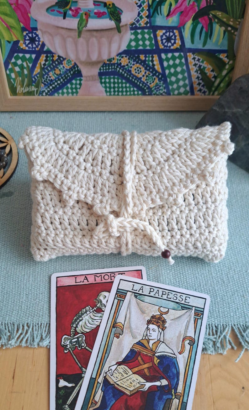 May include: A cream-coloured, hand-crocheted clutch bag with a flap and drawstring closure. Two tarot cards, "LA MORT" and "LA PAPESSE", are visible. A framed painting with birds and floral elements is in the background.