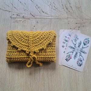 May include: Mustard yellow crochet clutch bag with a decorative flap and tie closure. Two tarot cards with floral and geometric designs are next to the bag. The text "Jaune moutarde" is at the bottom.