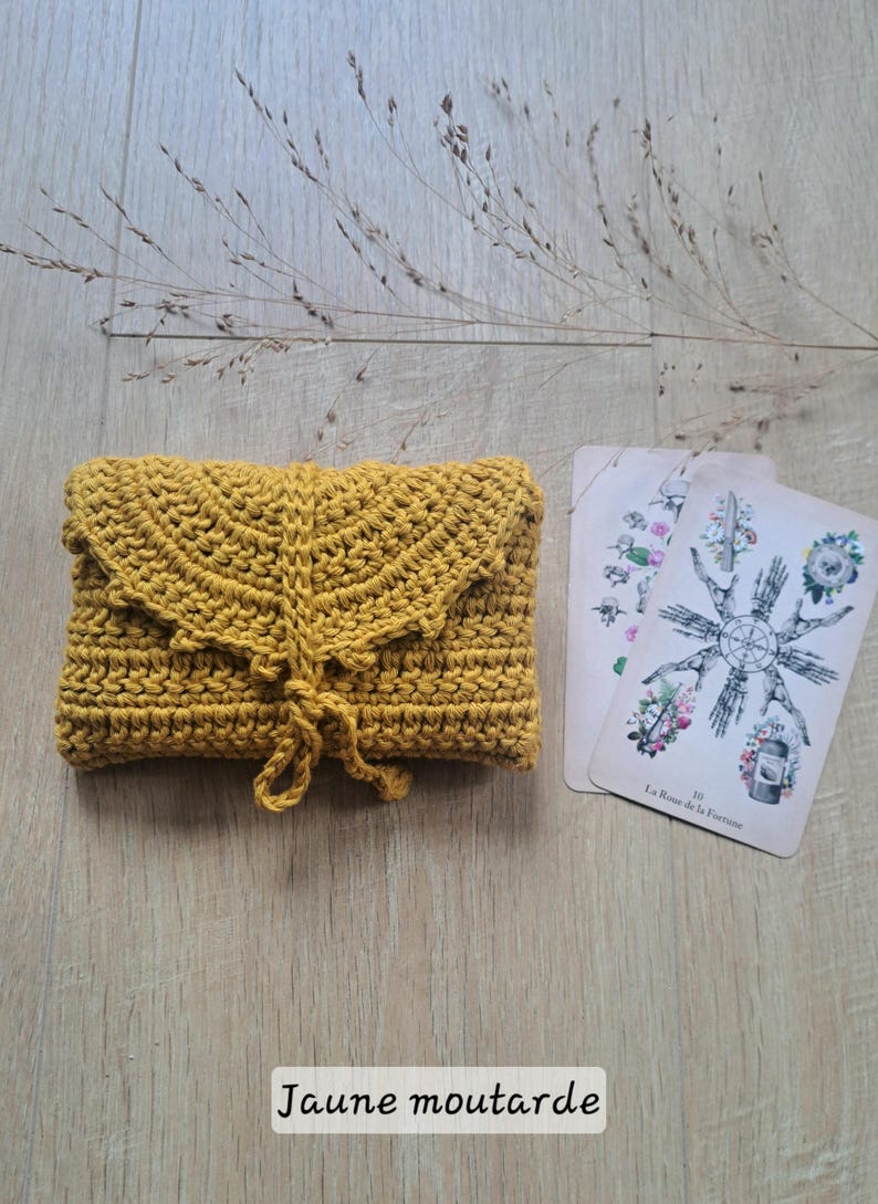 May include: Mustard yellow crochet clutch bag with a decorative flap and tie closure. Two tarot cards with floral and geometric designs are next to the bag. The text "Jaune moutarde" is at the bottom.