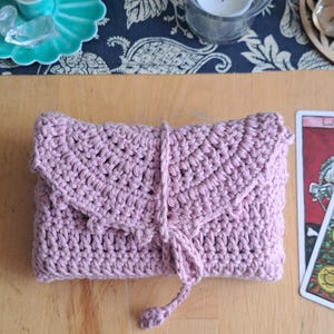 May include: A rose-pink crocheted clutch bag with a flap and tie closure. The handmade item is displayed on a wooden surface. A teal candle holder and a tarot card are visible in the background.
