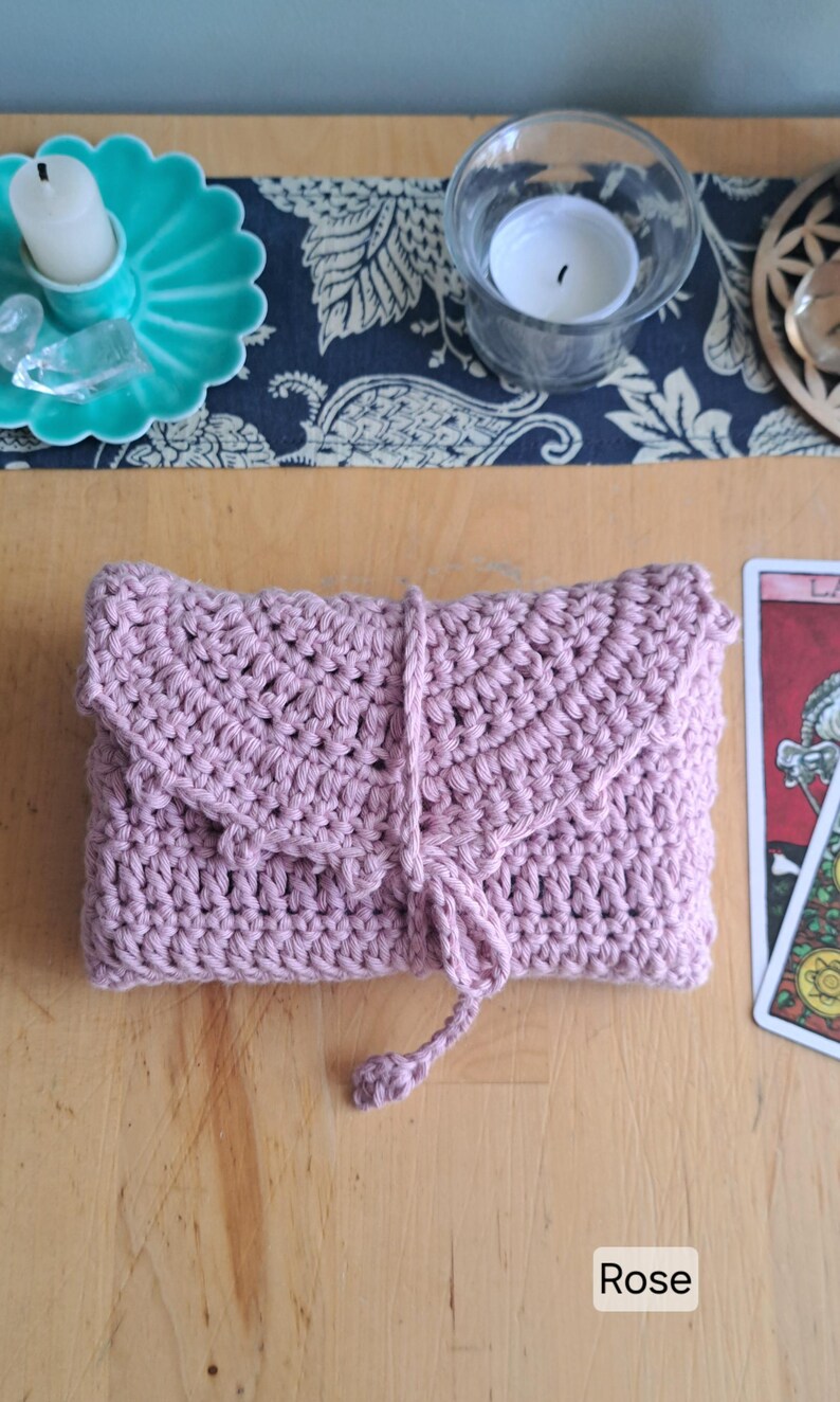 May include: A rose-pink crocheted clutch bag with a flap and tie closure. The handmade item is displayed on a wooden surface. A teal candle holder and a tarot card are visible in the background.