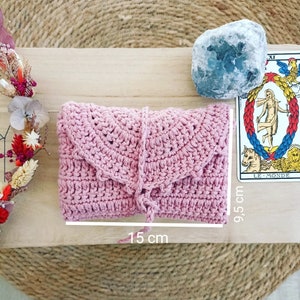 May include: A light pink crochet clutch, measuring 15 cm long and 9.5 cm high. The clutch has a flap closure and a matching tie. A tarot card and a blue crystal are next to the clutch.