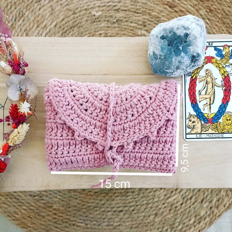 May include: A light pink crochet clutch, measuring 15 cm long and 9.5 cm high. The clutch has a flap closure and a matching tie. A tarot card and a blue crystal are next to the clutch.