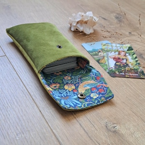 May include: A rectangular olive green suede pouch with a metal snap closure. The interior lining features a blue fabric with a multi-coloured floral and bird pattern. A deck of cards is partially inserted into the pouch, which is open.