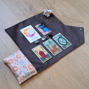 Könnte beinhalten: Eine dunkelbraune Tarot-Kartenmatte aus Stoff mit einer Umschlagtasche, die ein helles Muster aus orangen Sonnen und blauen Schlangen zeigt. Auf der Matte liegen vier Tarotkarten, drei kleine Kristalle und ein getrockneter weißer Blumenzweig.