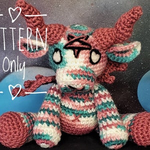 May include: Crocheted amigurumi toy of a pink, white, and teal striped cow with black eyes and a black nose. The toy is sitting on a dark surface. The text "PATTERN Only" is written in white on a black background.