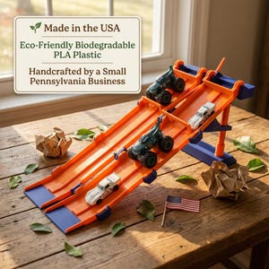 K&ouml;nnte beinhalten: Eine orangefarbene und blaue Spielzeug-Rennstrecke mit zwei Fahrspuren, auf der sich zwei Monstertrucks und zwei Rennwagen befinden. Die Rennstrecke besteht aus umweltfreundlichem, biologisch abbaubarem PLA-Kunststoff und wird in Pennsylvania handgefertigt. Eine kleine amerikanische Flagge ist auf der Holzoberfl&auml;che platziert.