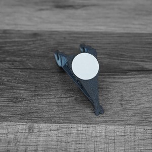Puede incluir: Un objeto gris oscuro impreso en 3D con una pegatina circular blanca. El objeto tiene una forma &uacute;nica, posiblemente un clip o soporte, con un dise&ntilde;o en forma de aleta. El art&iacute;culo est&aacute; sobre un fondo de madera.